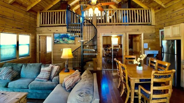 Un interior de cabaña con una escalera de madera, un comedor y una sala de estar con sofá y lámpara.