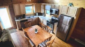 Interior de una cabaña de madera con una mesa de comedor, sillas, una cocina equipada con refrigerador, microondas y estufa.