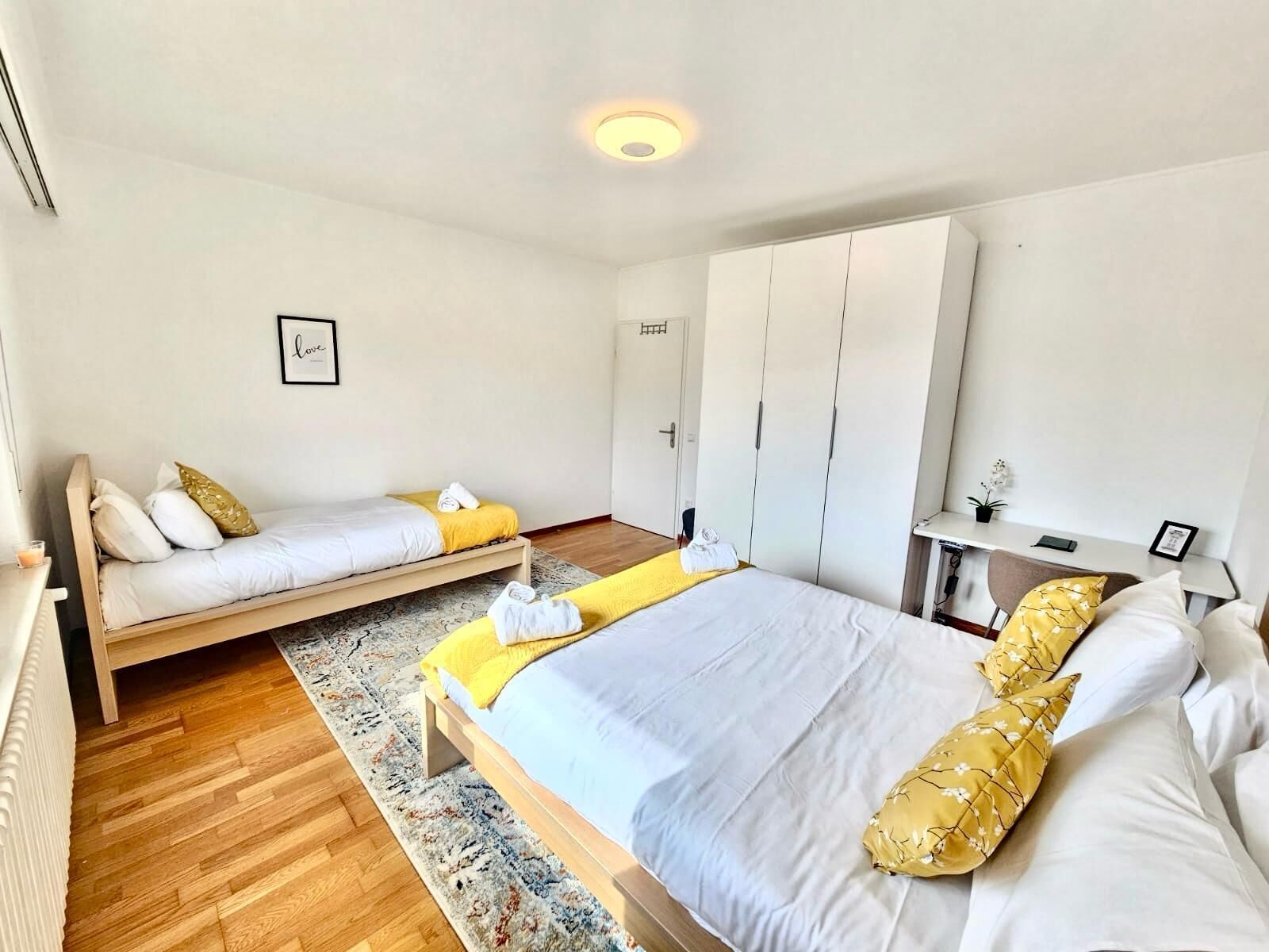 A modern bedroom with a large bed, a smaller bed, a wardrobe, and a desk with a chair.