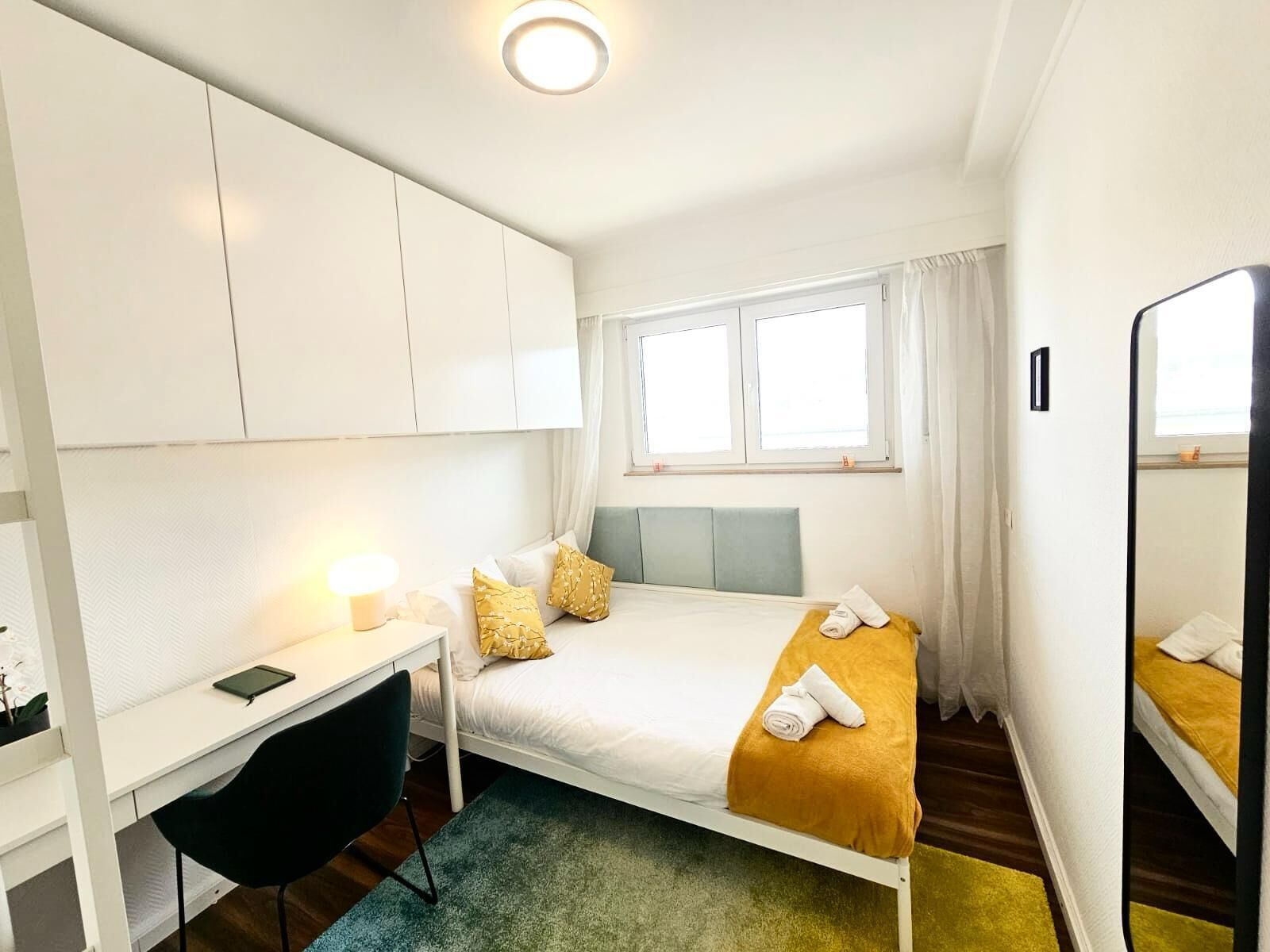 Habitación de hotel pequeña con una cama individual, un escritorio con silla, una mesita de noche, un espejo y una ventana con cortinas.