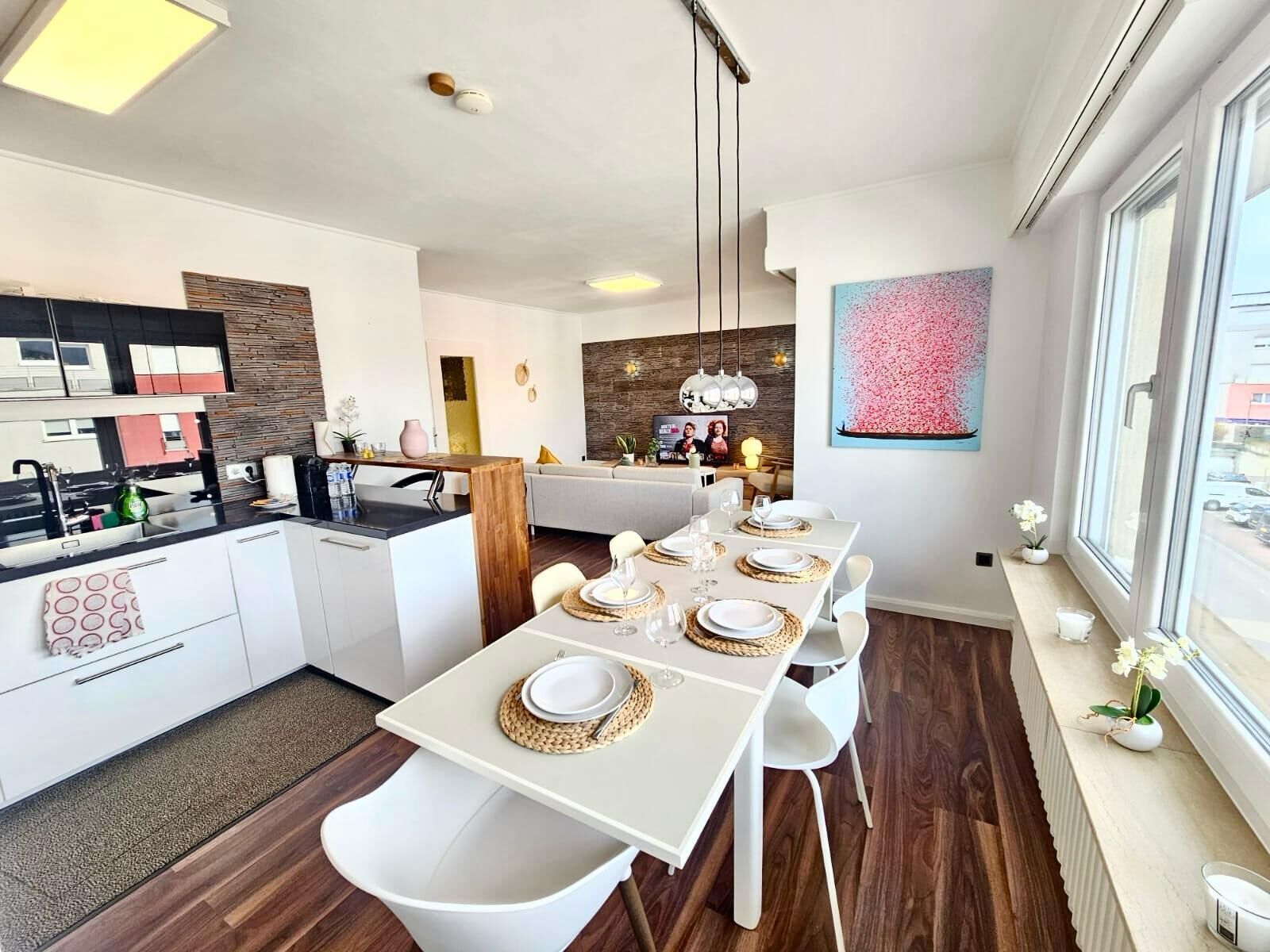 A modern kitchen with a dining area, featuring a long table set for four, a built-in oven, and a large window with a view.