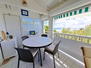 A covered outdoor seating area with a round table and chairs, a small white cabinet, and a view of the ocean.