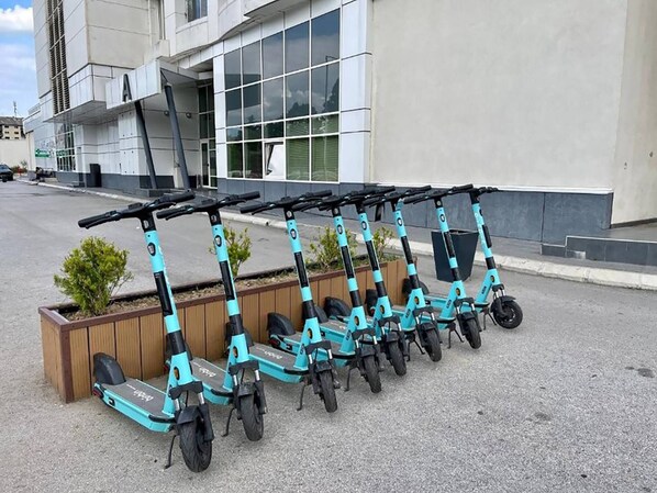 A row of teal electric scooters parked in front of a modern building with large windows and a wooden planter with small plants.