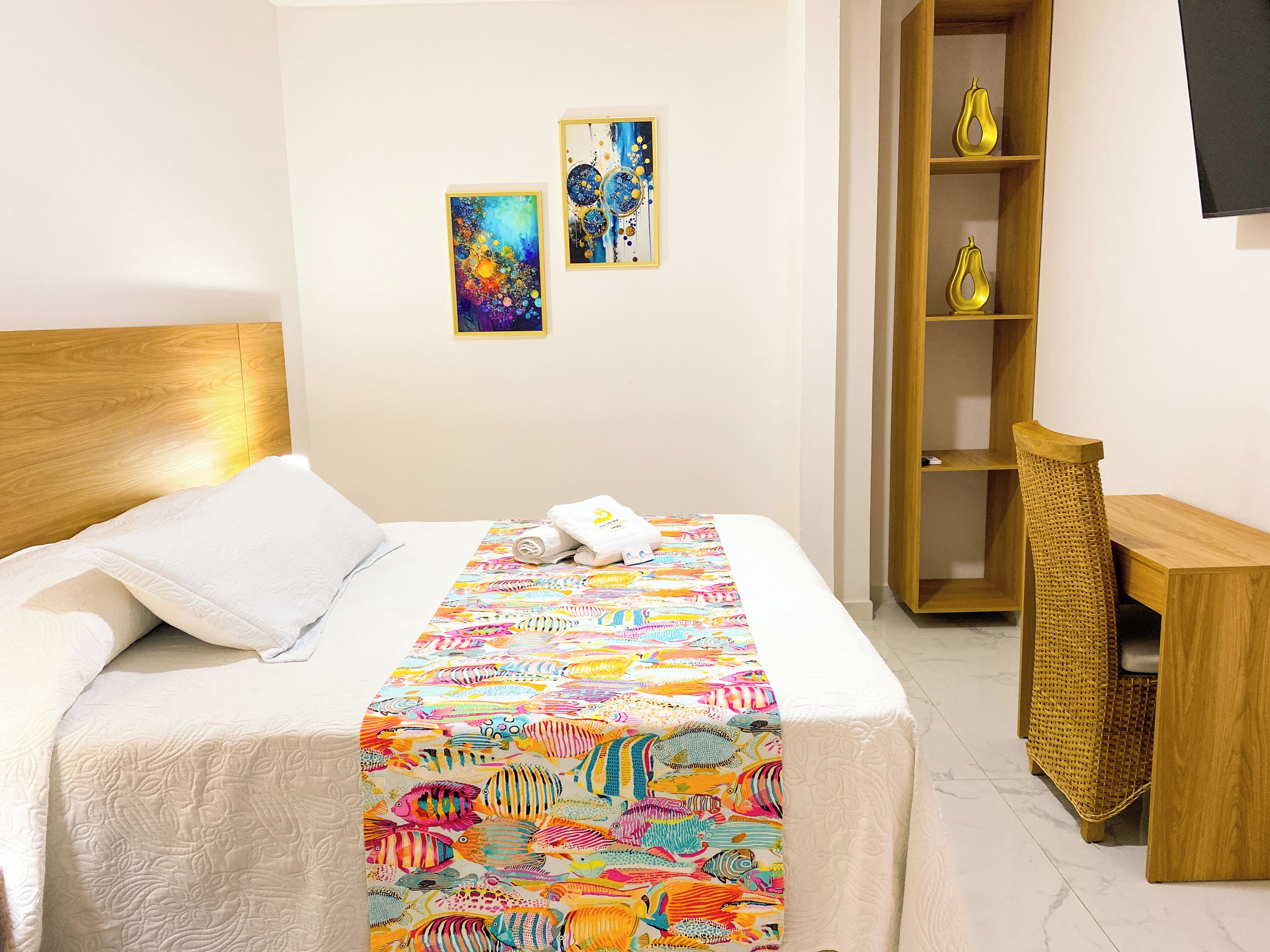 A hotel room with a bed, a wooden headboard, a colorful bedspread, two framed artworks, a wooden side table, and a wicker chair.