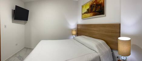 A hotel room with a bed, a mounted TV, a painting on the wall, and a bedside lamp.