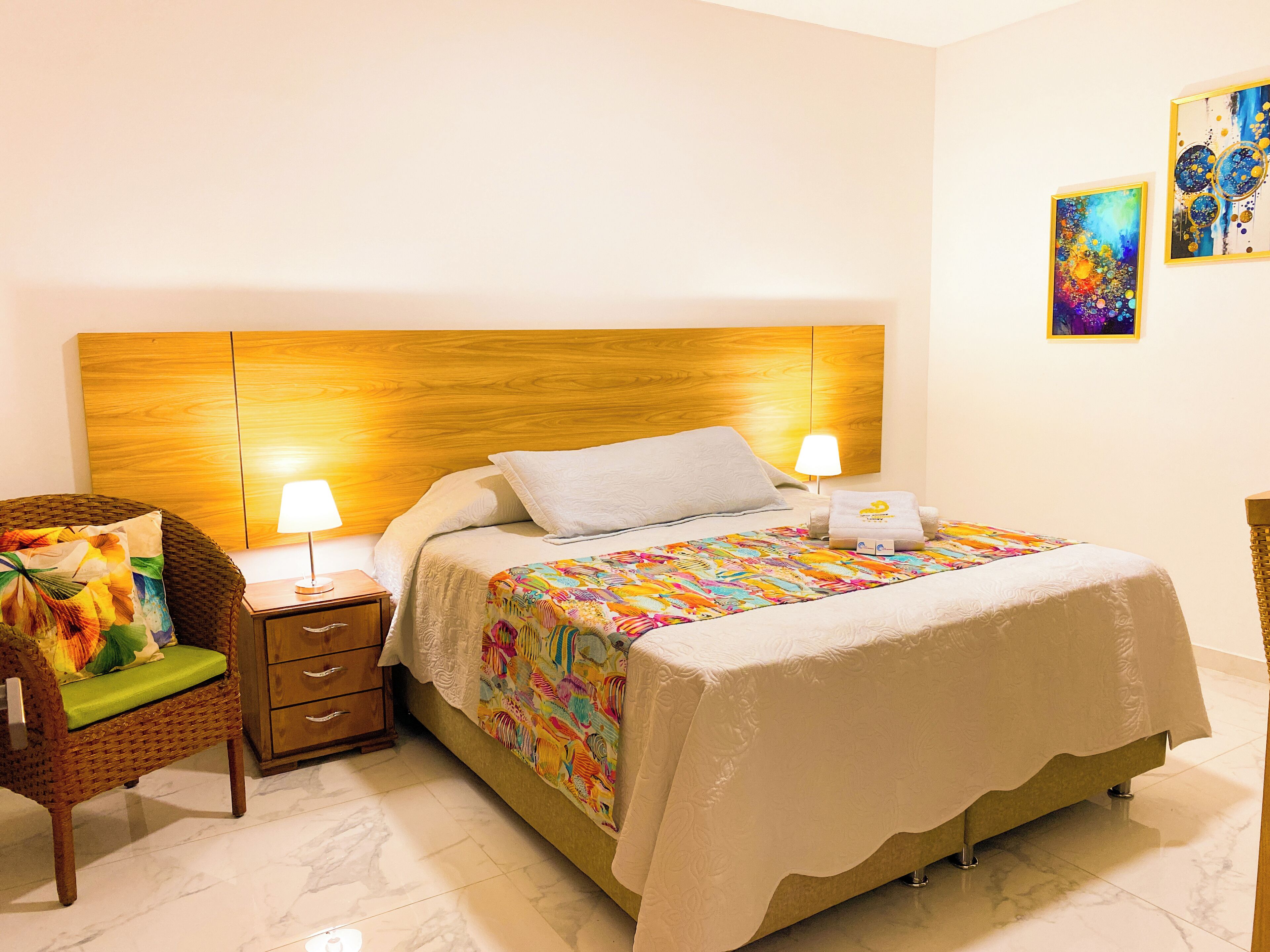 A neatly arranged hotel room with a wooden headboard, a bed with a colorful quilt, a wicker chair, and two bedside lamps.