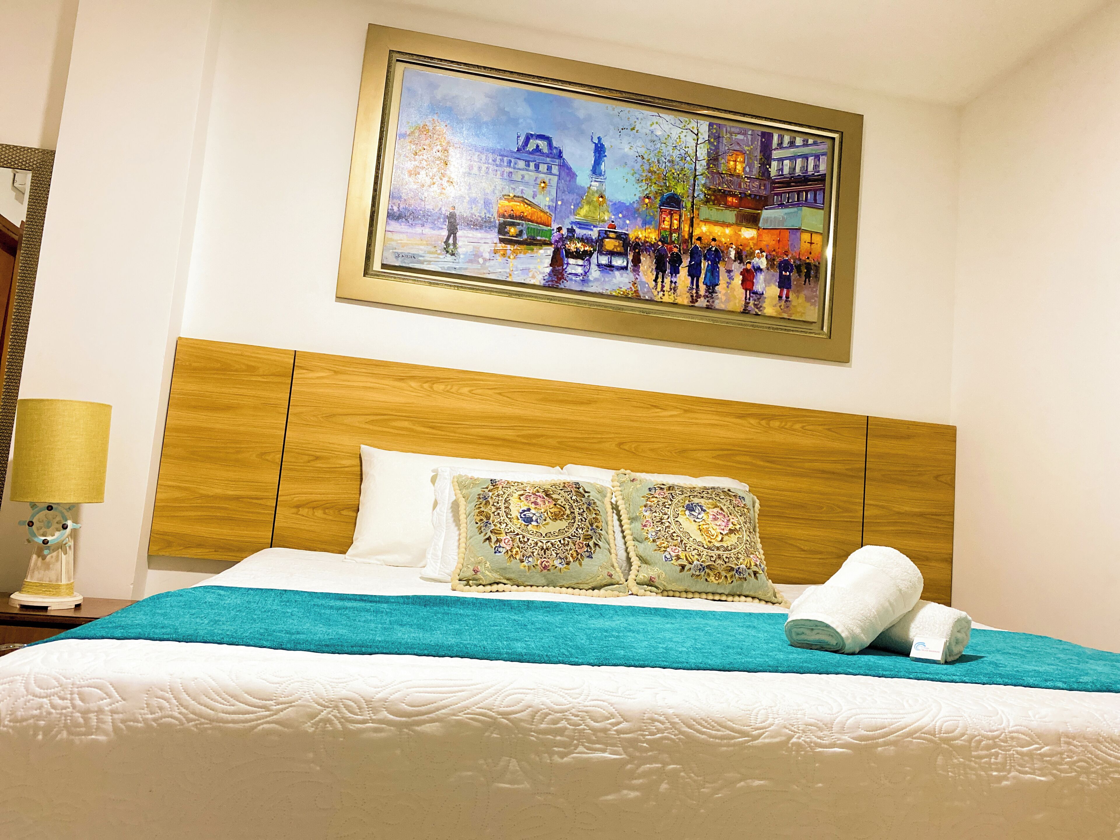 Ein Hotelzimmer mit einem hölzernen Kopfteil, einem Bett mit weißer Bettwäsche, zwei Zierkissen und einem Stadtbild über dem Bett.