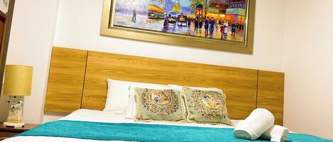 A hotel room with a wooden headboard, a bed with white bedding, two decorative pillows, and a painting of a city scene above the bed.