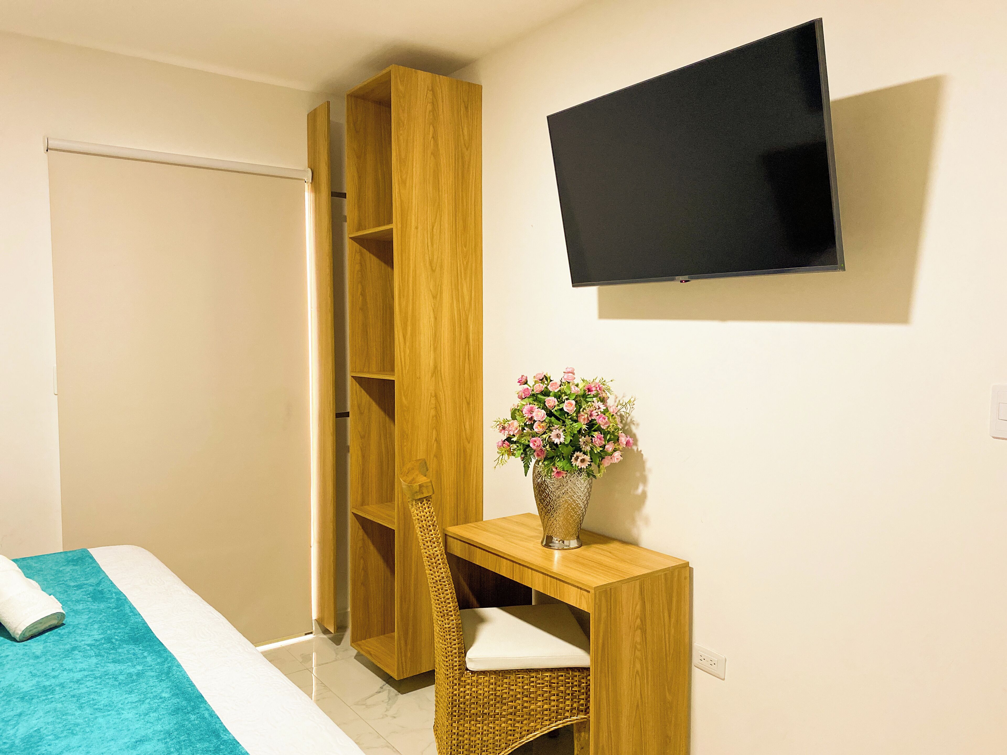 Une chambre à coucher avec un lit, une armoire en bois, un bureau agrémenté d’un vase de fleurs et un téléviseur fixé au mur.