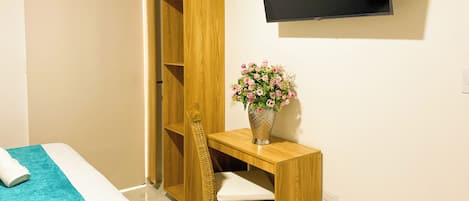 A bedroom with a bed, a wooden wardrobe, a desk with a vase of flowers, and a mounted TV.