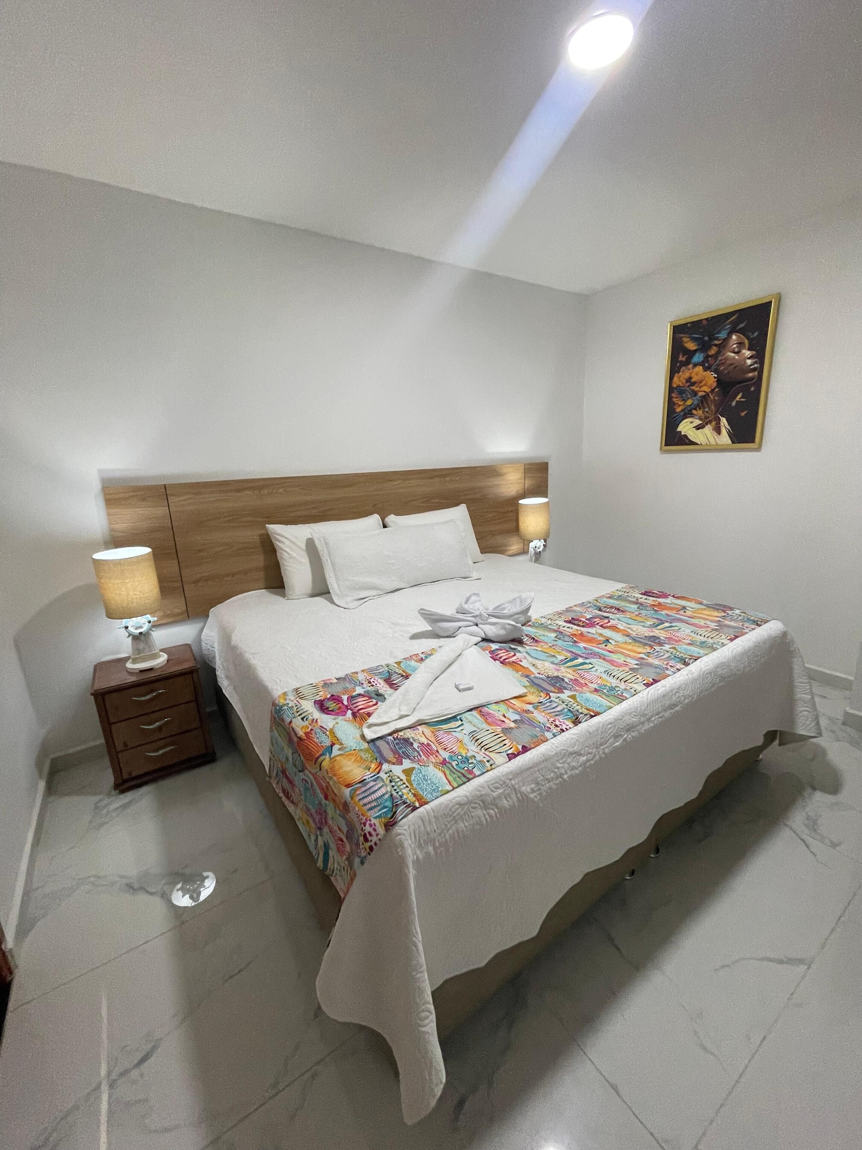 A neatly made bed with a colorful bedspread, two bedside lamps, a nightstand with a lamp, and a painting on the wall.