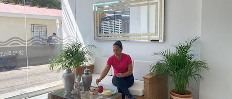 Eine Person sitzt auf einem weißen Sofa in einem Raum mit einem Glastisch, zwei Blumentöpfen und einem großen Spiegel an der Wand.