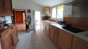 A kitchen with wooden cabinets, a stainless steel range hood, a window with a yellow curtain, and a tiled floor. - Charming apartment in a 17th century farmhouse, sleeps 4 (LE CHATEAU DOLERON)