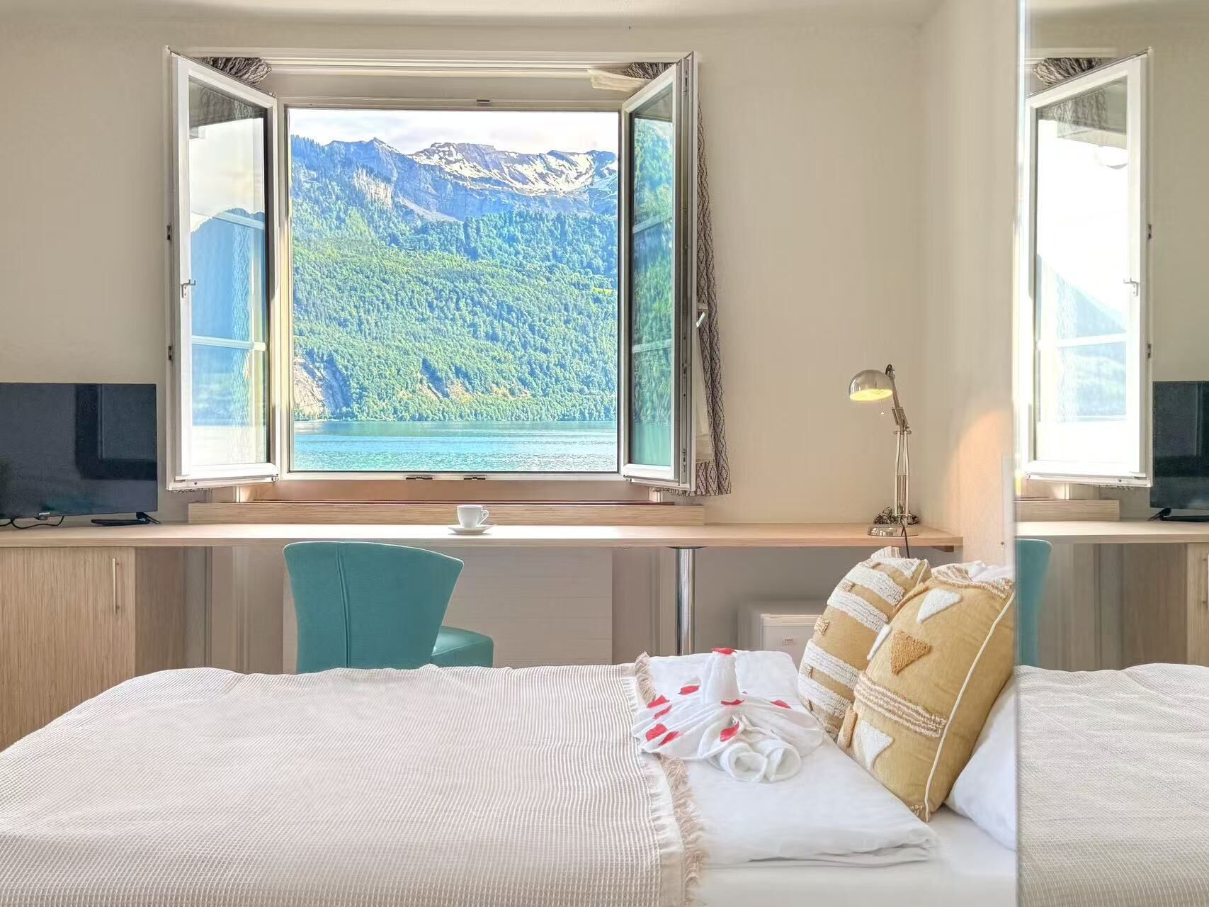 Une chambre dotée d’une grande fenêtre donnant sur les montagnes, d’un bureau avec un ordinateur et d’un lit recouvert d’une couette blanche et agrémenté de coussins décoratifs.