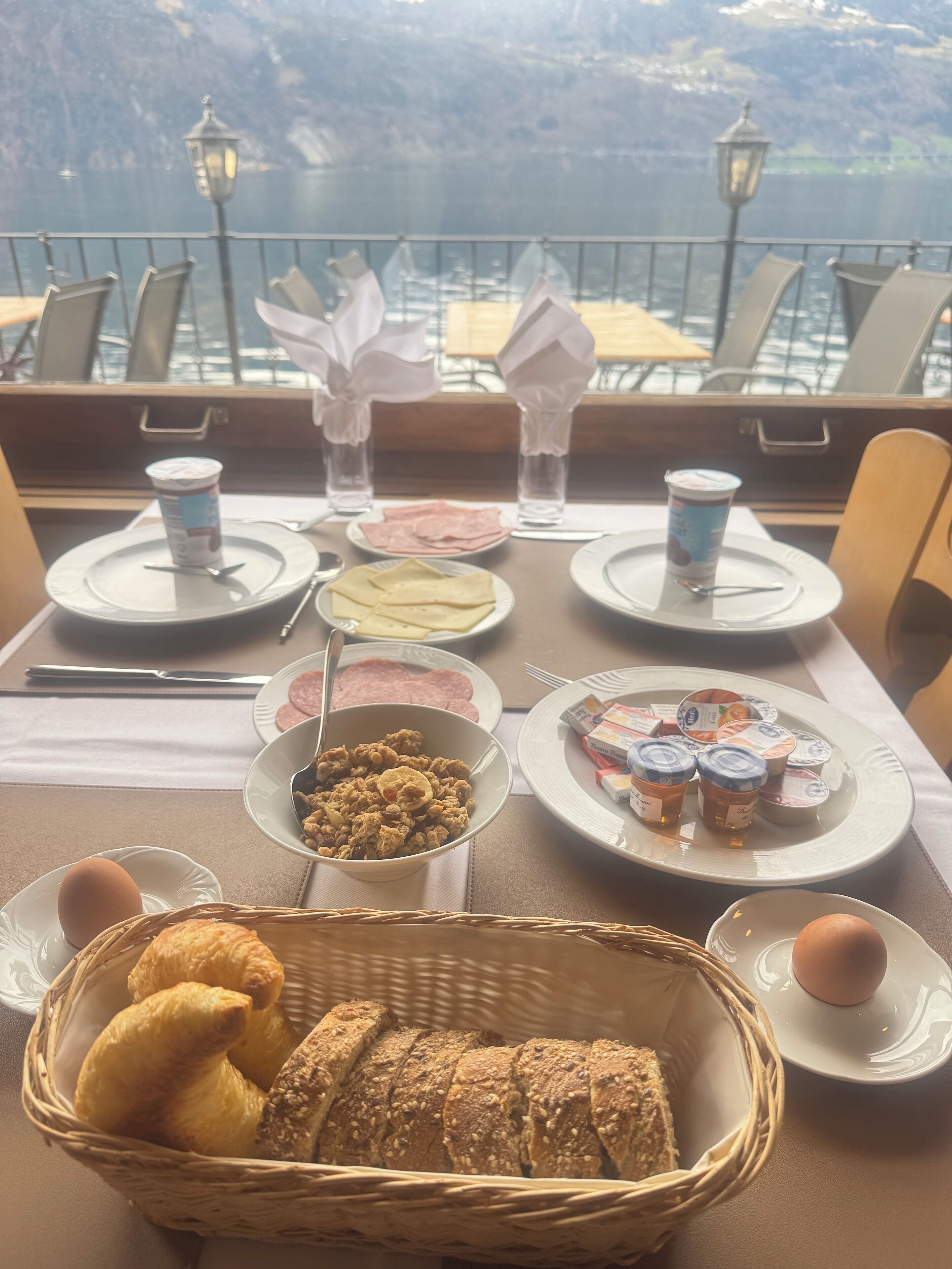 A breakfast setting with croissants, bread, eggs, and yogurt cups on a table with a view of a lake.