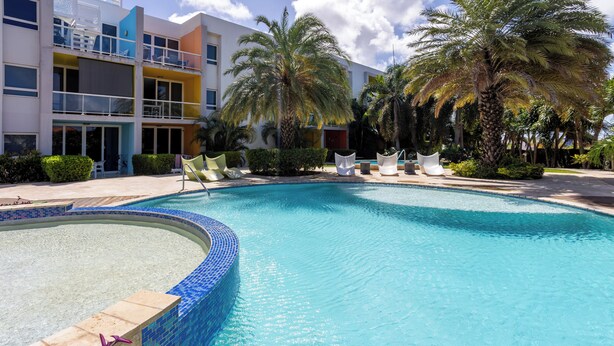 Una piscina a forma curva, circondata da poltrone a sdraio e palme, con un edificio a più piani sullo sfondo.