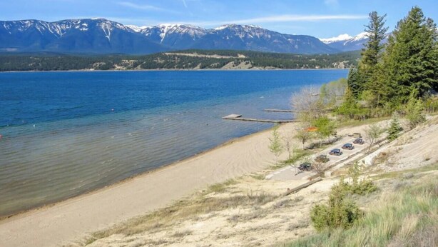 A lakeside view with a sandy beach, a dock, and mountains in the background.
