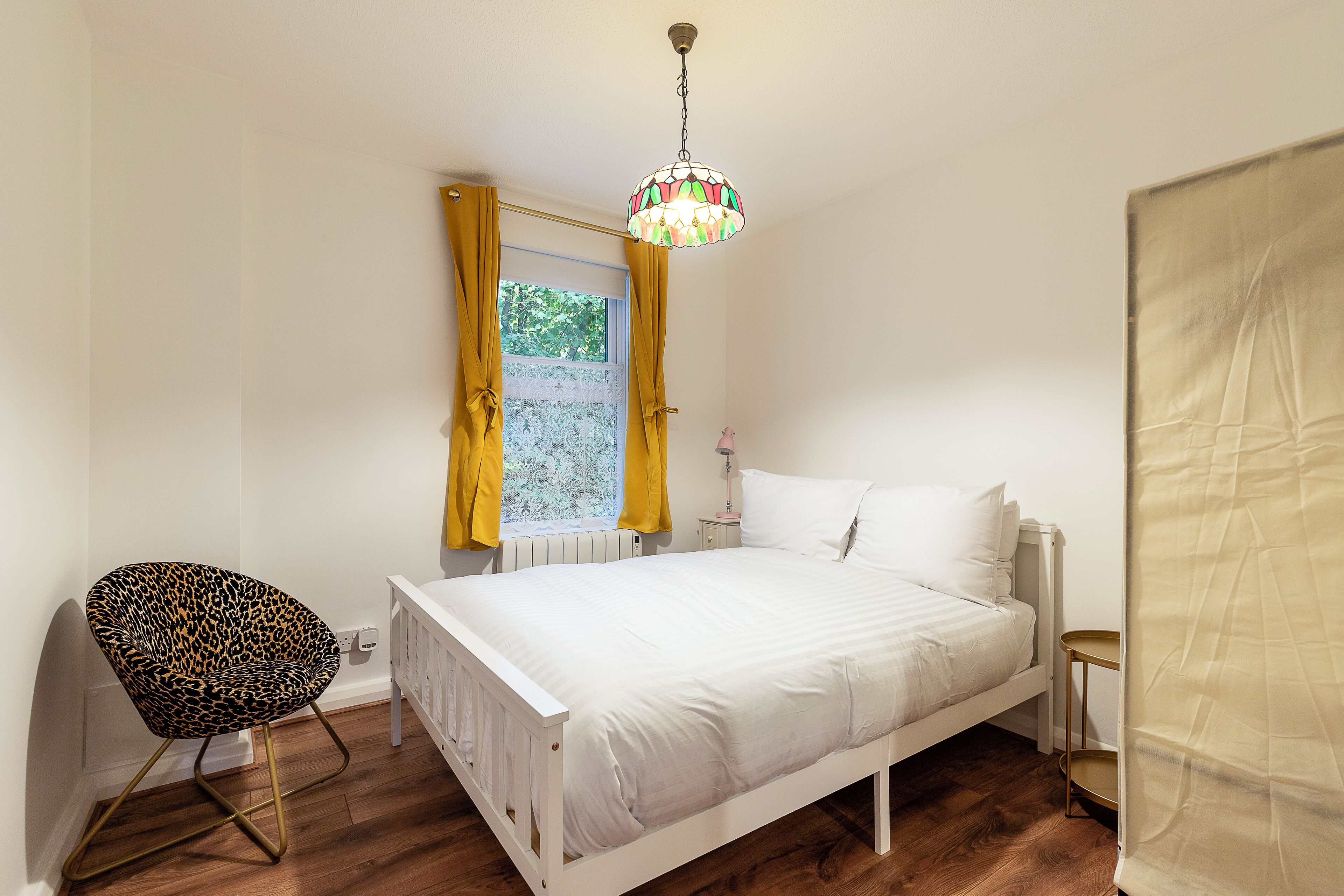 Un dormitorio con una cama blanca, un sillón estampado de leopardo, una mesita de noche pequeña y una ventana con cortinas amarillas.
