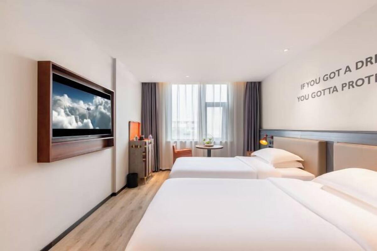Ein Hotelzimmer mit zwei Betten, einem an der Wand befestigten Fernseher, einem kleinen Tisch mit einer Lampe und einem Fenster mit Vorhängen.