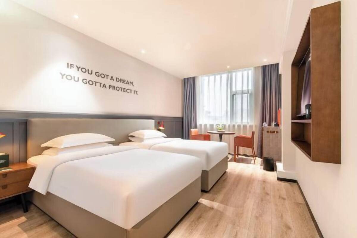 Ein Hotelzimmer mit zwei Betten, einem Schreibtisch, einem Stuhl und einem großen Fenster mit Vorhängen.