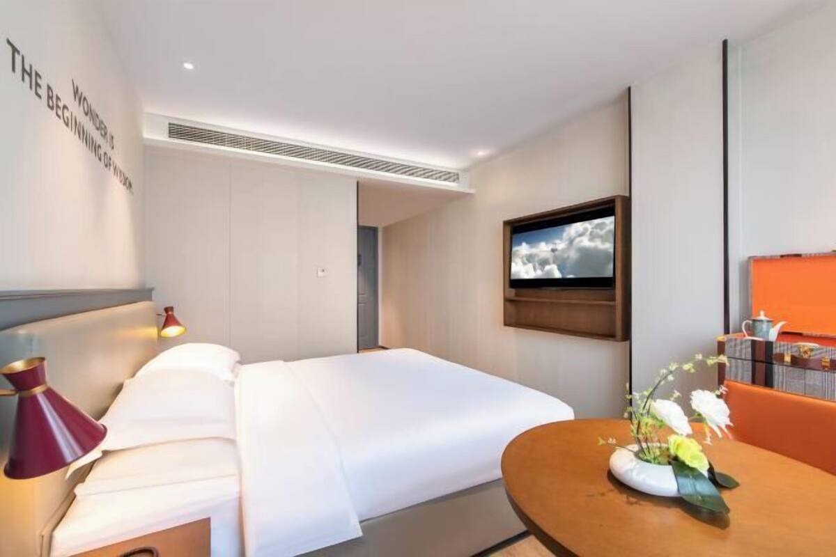 Ein Hotelzimmer mit Bett, einem an der Wand montierten Fernseher, einem Tisch mit Blumenvase und einer Wand mit einem Schild.