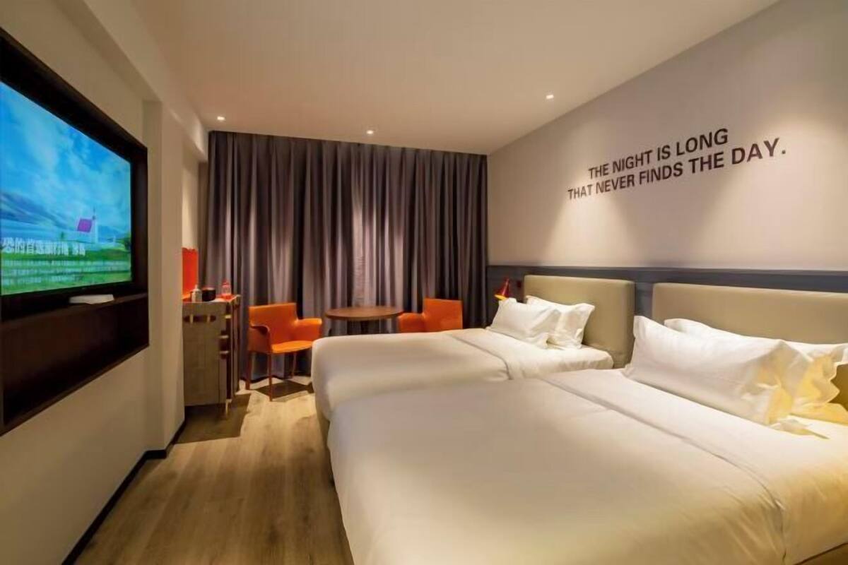Ein Hotelzimmer mit einem großen Bett, einem Fernseher, einem Schreibtisch und einem Stuhl. An der Wand ist ein Zitat zu sehen.