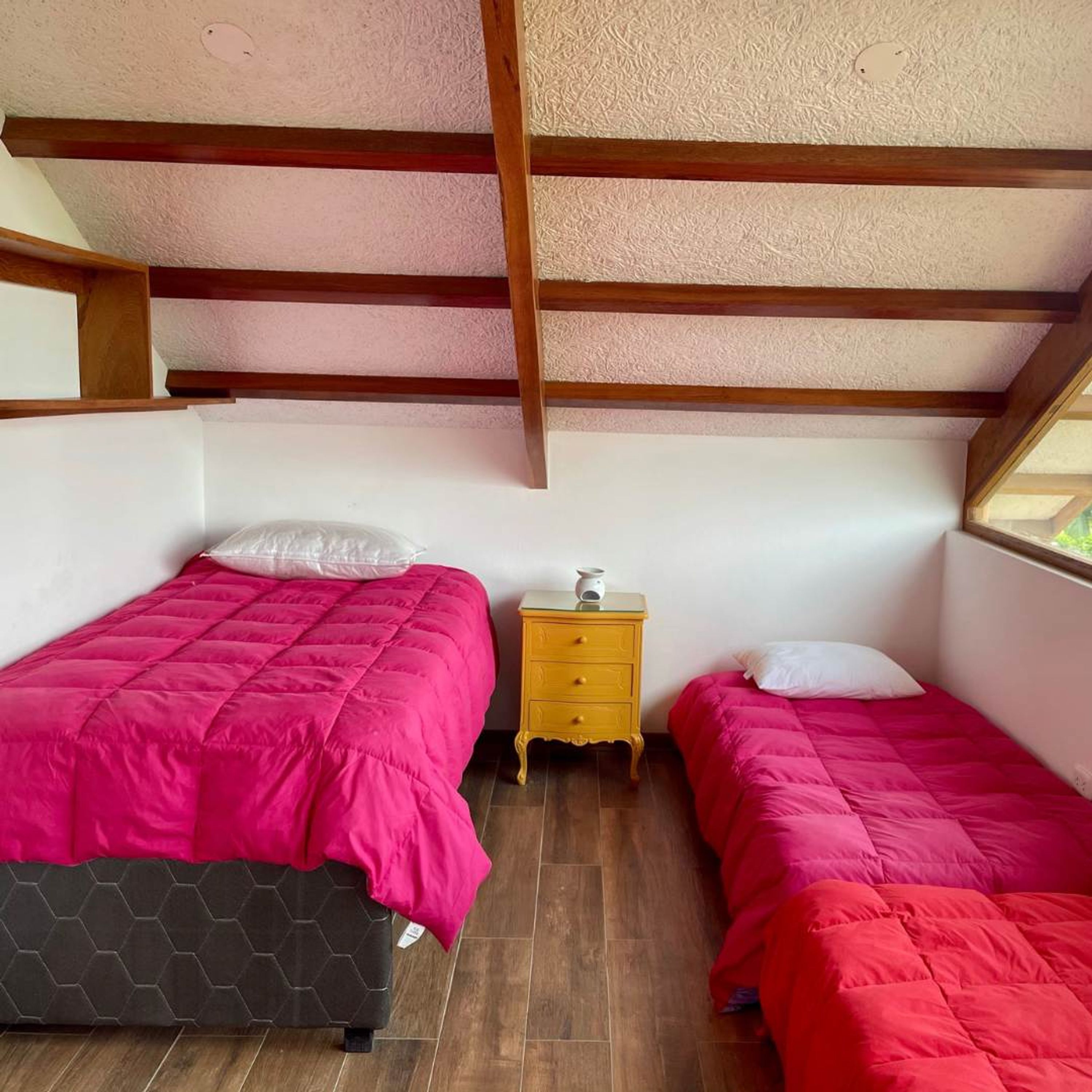 A small, well-lit room with two beds, a wooden nightstand, and a window.
