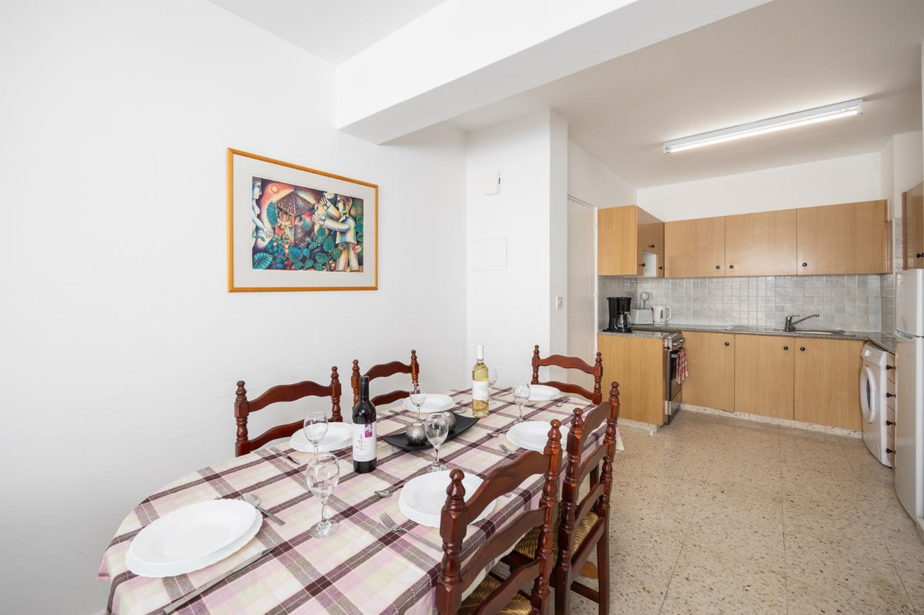 A dining area with a table set for four, wooden chairs, a painting on the wall, and a kitchen in the background.