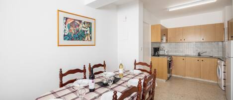 A dining area with a table set for four, wooden chairs, a painting on the wall, and a kitchen in the background.