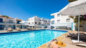 A swimming pool with lounge chairs and umbrellas, surrounded by white buildings with balconies.