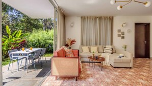 A living room with a sofa, a dining table with chairs, a coffee table, and a large window with curtains. - Stylish 2BR w Pool Near Candolim and Coco Beach (Verem)
