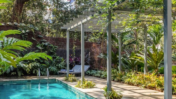 A rectangular swimming pool with clear blue water, surrounded by lush greenery and a pergola structure. - Stylish 2BR w Pool Near Candolim and Coco Beach (Verem)