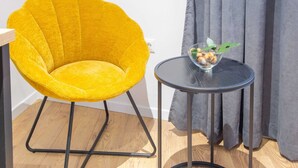 Una silla amarilla con estructura metálica negra, una mesita redonda con un bol de vidrio que contiene aperitivos, y una cortina gris al fondo.