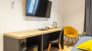 Una habitación de hotel moderna con un escritorio de madera, una silla negra, un sillón amarillo, una cama con almohada amarilla, un televisor de pantalla plana y una mesita con tetera y tazas.