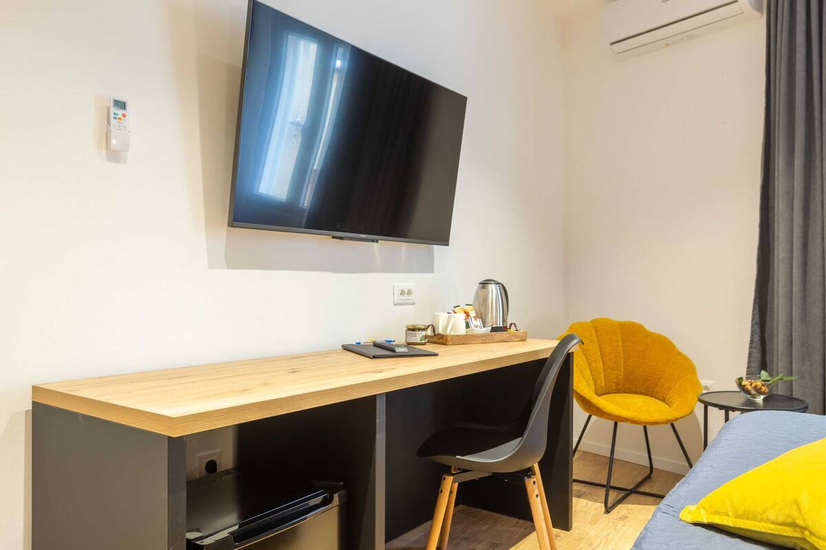 Una habitación de hotel moderna con un escritorio de madera, una silla negra, un sillón amarillo, una cama con almohada amarilla, un televisor de pantalla plana y una mesita con tetera y tazas.