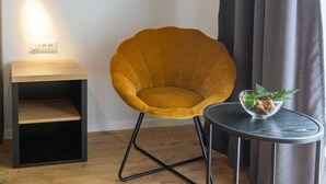 Una silla de color mostaza con estructura metálica negra, una mesita de madera con un cuenco de vidrio y hojas verdes, y un enchufe de pared.