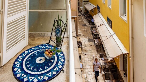 Una estrecha calle con una mesa y sillas de diseño azul y blanco, una persona caminando y edificios con balcones y toldos.