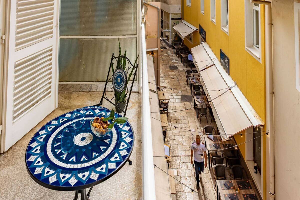 Una estrecha calle con una mesa y sillas de diseño azul y blanco, una persona caminando y edificios con balcones y toldos.