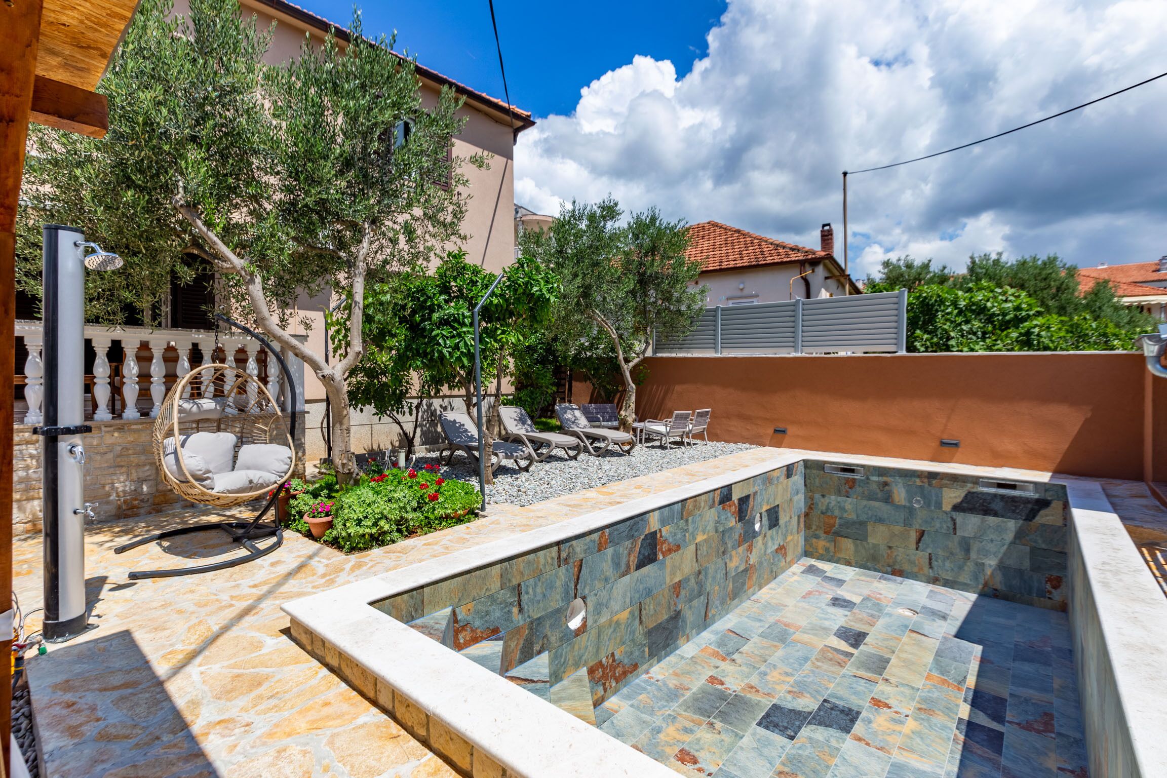 A swimming pool with stone tiles, surrounded by a stone wall and a wooden pergola with chairs.