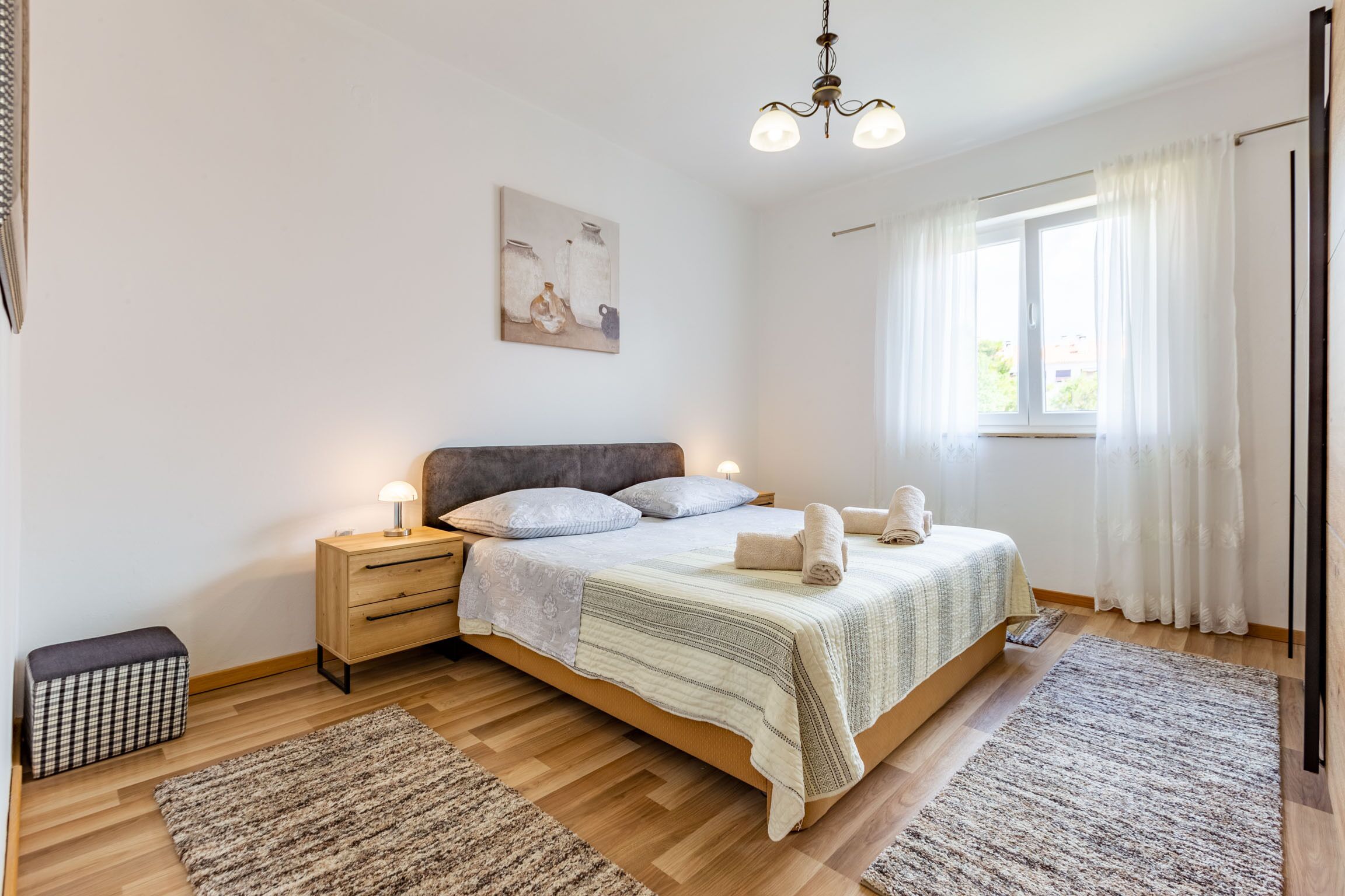 Une chambre à coucher avec un lit, des tables de chevet, une commode, une lampe, un tableau, une fenêtre avec des rideaux et un tapis.