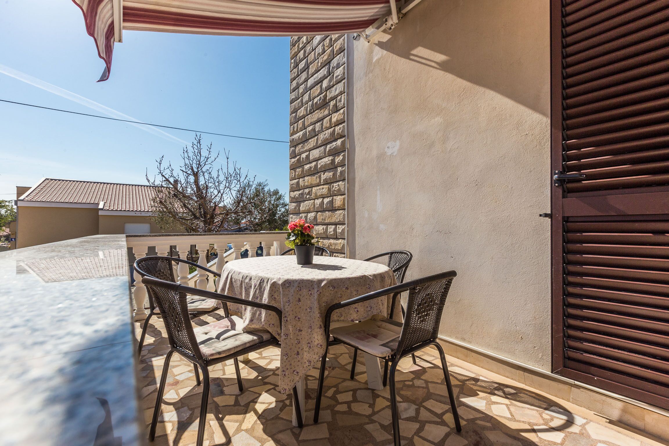 A patio with a round table and four chairs, a potted plant with flowers, and a brown shutter door.