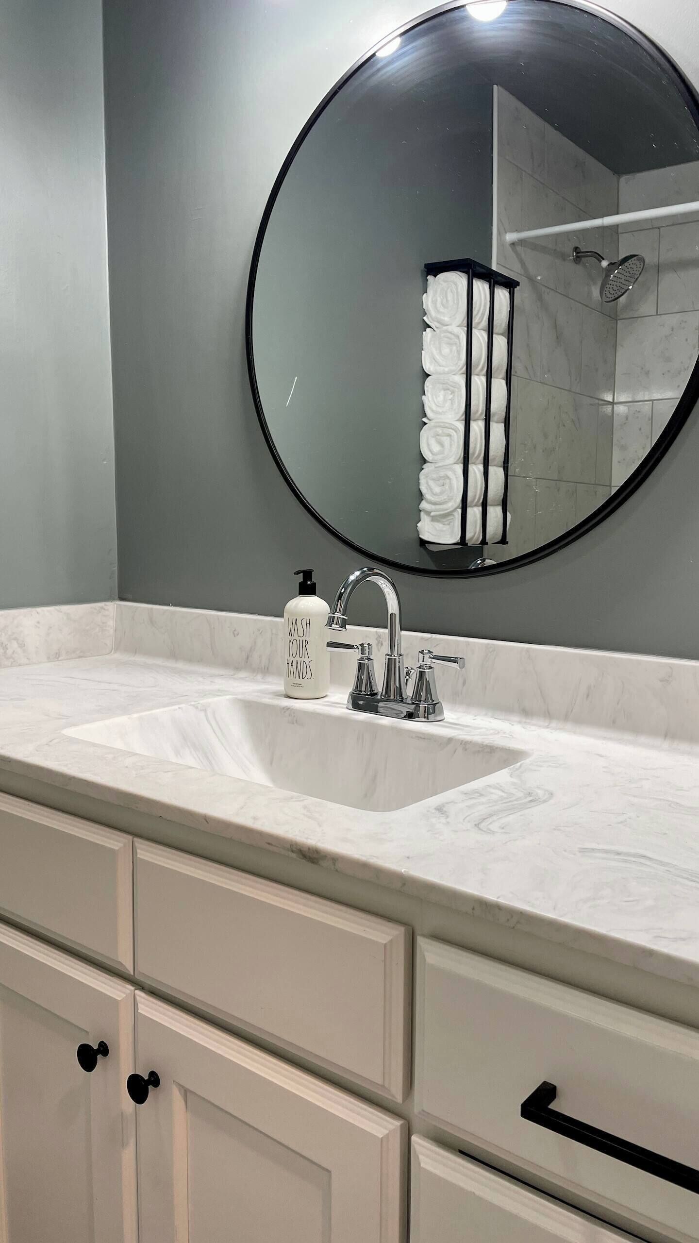 A bathroom with a round mirror, two sinks, a towel rack, and a bottle of hand soap.