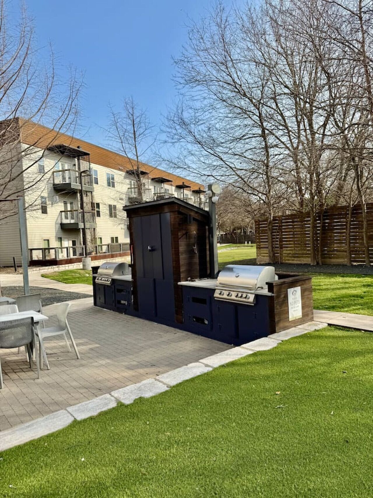 Un espace barbecue extérieur équipé d’un gril et de sièges, entouré d’une pelouse verdoyante et d’une clôture en bois.