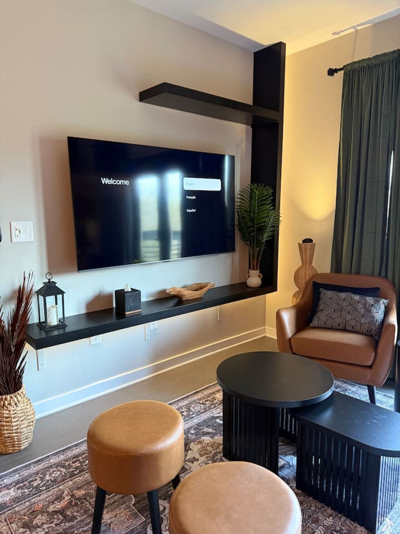 Un salon moderne avec un grand téléviseur à écran plat fixé au mur, un canapé en cuir marron, deux poufs ronds, une table basse noire et une plante décorative.
