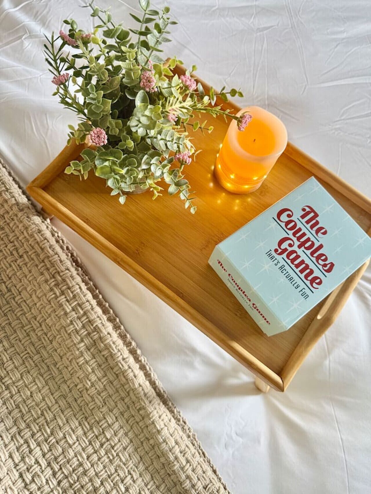 Una bandeja de madera con un libro titulado 'The Couples Game' y una vela sobre una cama.