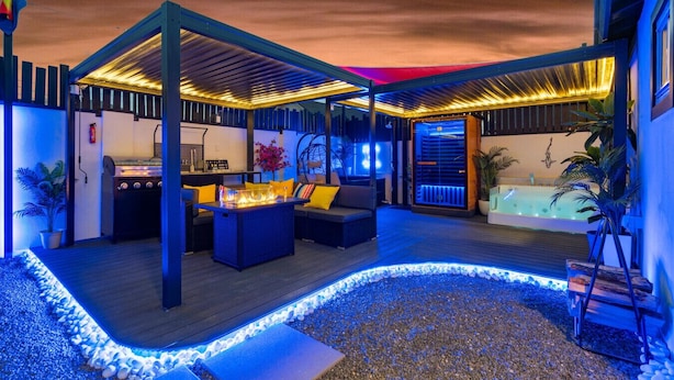 Un patio al aire libre con una parrilla, asientos y un jacuzzi, iluminado con luces ambientales.