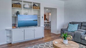 Een moderne woonkamer met een flatscreen televisie op een witte kast, een grijze bank met decoratieve kussens en een ronde houten salontafel met een plant.