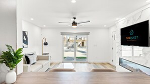 A modern living room with a wooden dining table, a ceiling fan, a flat-screen TV, and a large window with a view of the outside. - Ocean Breeze (Panama City Beach)
