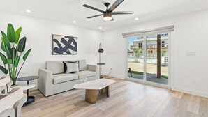 A spacious living room with a ceiling fan, a sofa, a coffee table, and a large window with a view of a residential area. - Ocean Breeze (Panama City Beach)