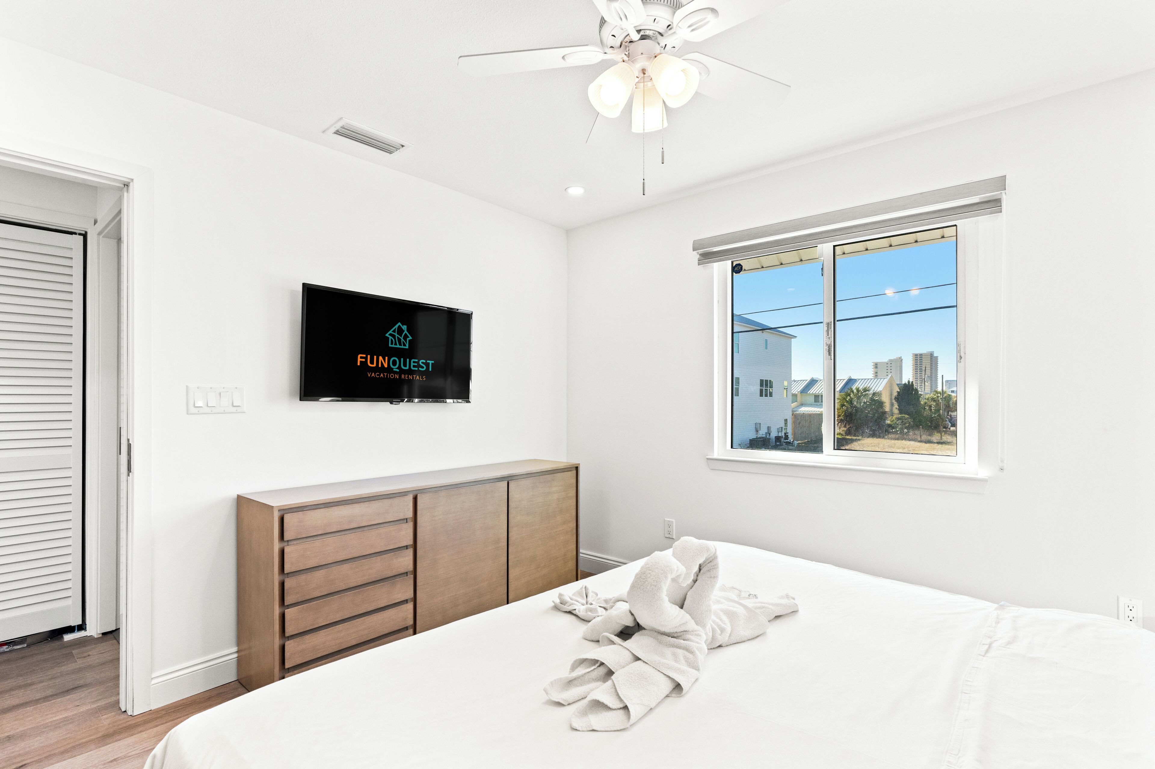 A modern hotel room with a bed, a TV mounted on the wall, a window with a city view, and a ceiling fan.